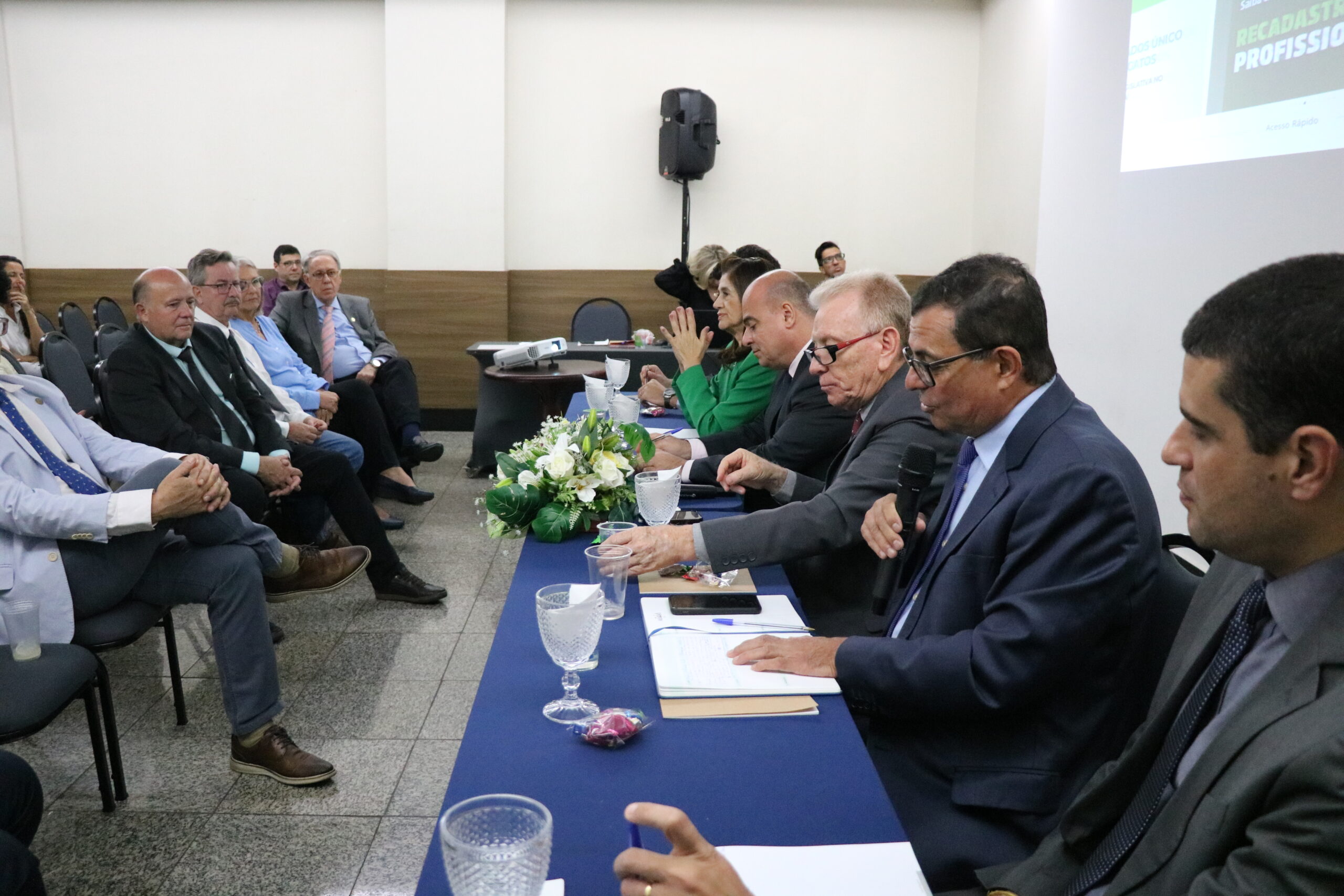 A imagem mostra os seis representantes do CRMV-SP, uma mulher de blazer verde, e cinco homens de terno escuro, sentados à mesa, sendo observados pelas pessoas presentes ao evento.