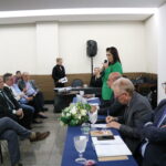 A imagem traz uma das representantes do CRMV-SP, de blazer verde, falando ao microfone para os presentes, os outros integrantes da mesa permanecem sentados.