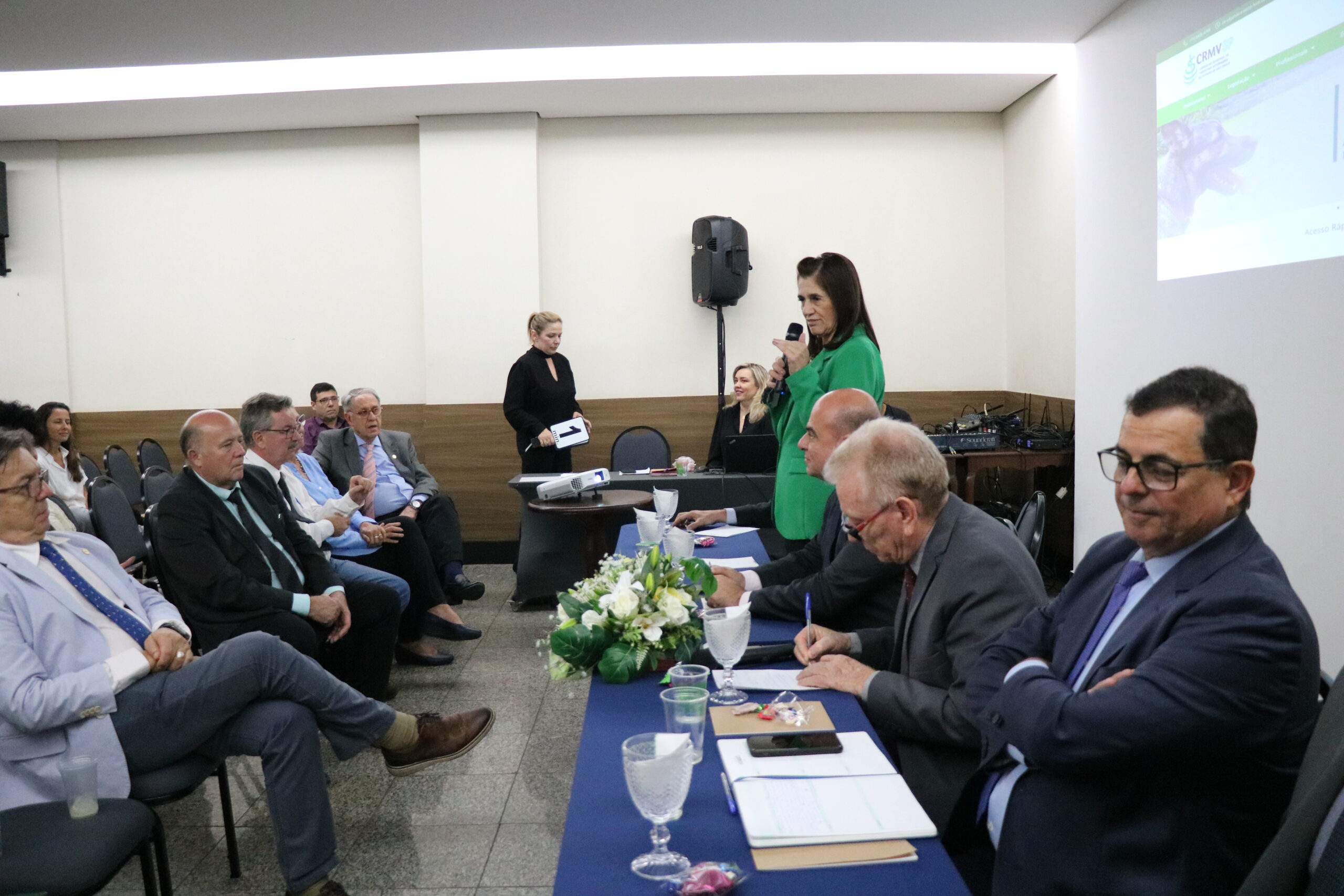 A imagem traz uma das representantes do CRMV-SP, de blazer verde, falando ao microfone para os presentes, os outros integrantes da mesa permanecem sentados.