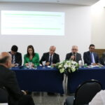 A imagem traz seis representantes do CRMV-SP, uma mulher de blazer verde, e cinco homens de terno escuro, sentados à mesa, lado a lado. O presidente do Conselho fala ao microfone para pessoas presentes no auditório.