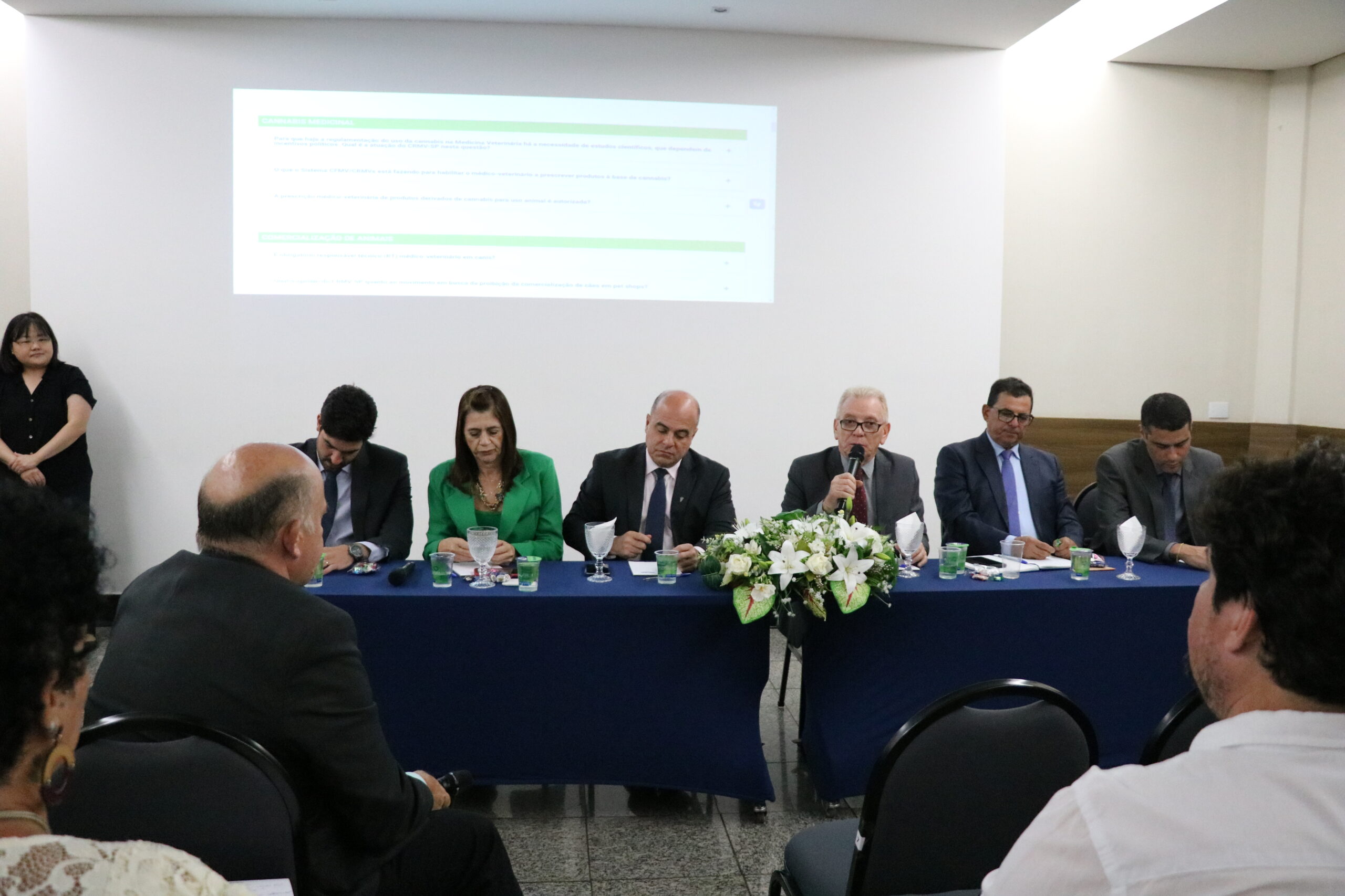 A imagem traz seis representantes do CRMV-SP, uma mulher de blazer verde, e cinco homens de terno escuro, sentados à mesa, lado a lado. O presidente do Conselho fala ao microfone para pessoas presentes no auditório.