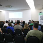 A imagem traz as pessoas presentes ao evento sentadas observando a mesa ao fundo, com os representantes do CRMV-SP. O presidente do CRMV-SP, de terno escuro, de óculos, com o microfone na mão, fala sentado à mesa.