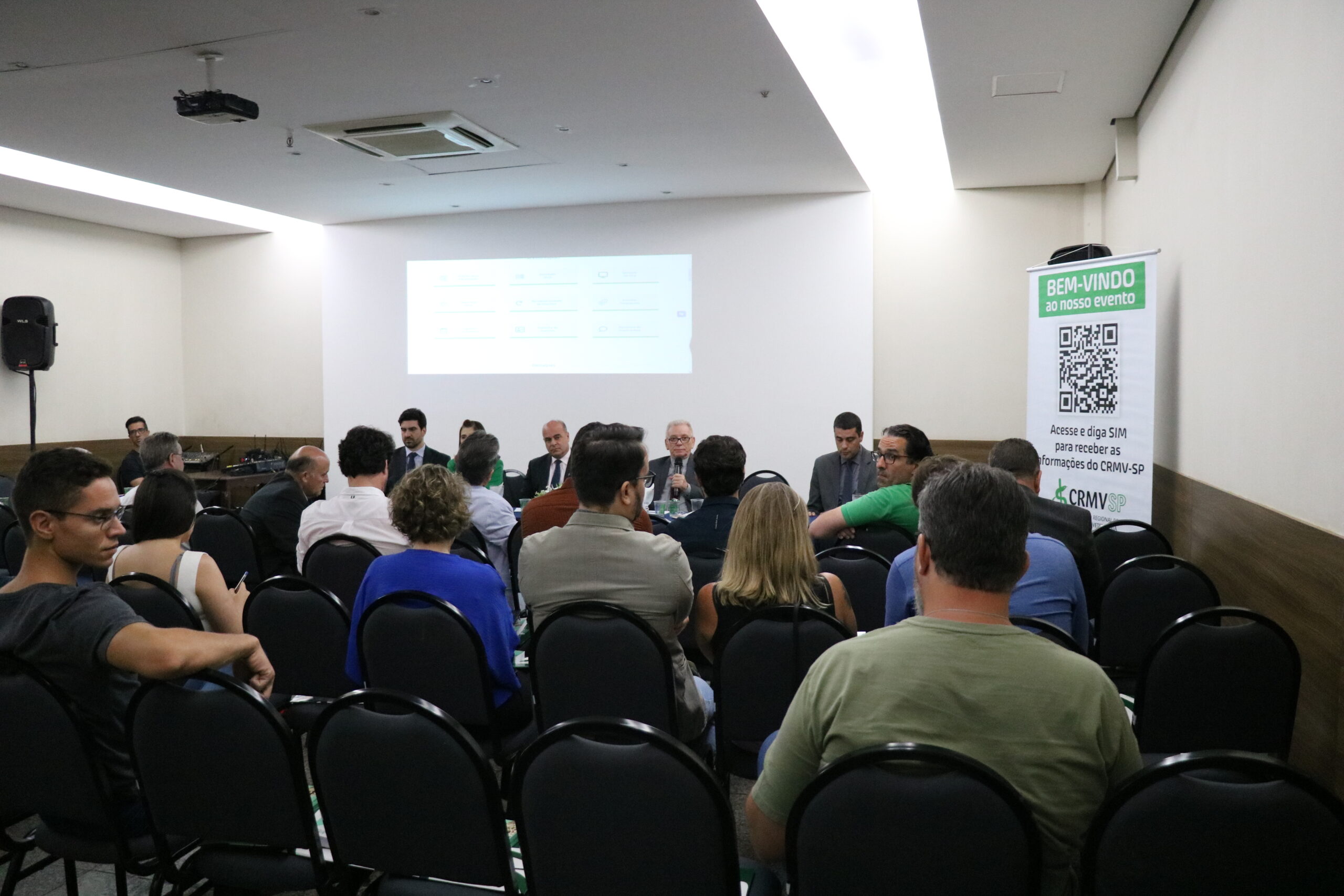 A imagem traz as pessoas presentes ao evento sentadas observando a mesa ao fundo, com os representantes do CRMV-SP. O presidente do CRMV-SP, de terno escuro, de óculos, com o microfone na mão, fala sentado à mesa.