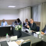 A imagem aproximada, traz seis pessoas, duas mulheres e quatro homens, sendo três deles de óculos. Eles estão sentados à mesa, um deles está falando ao microfone.