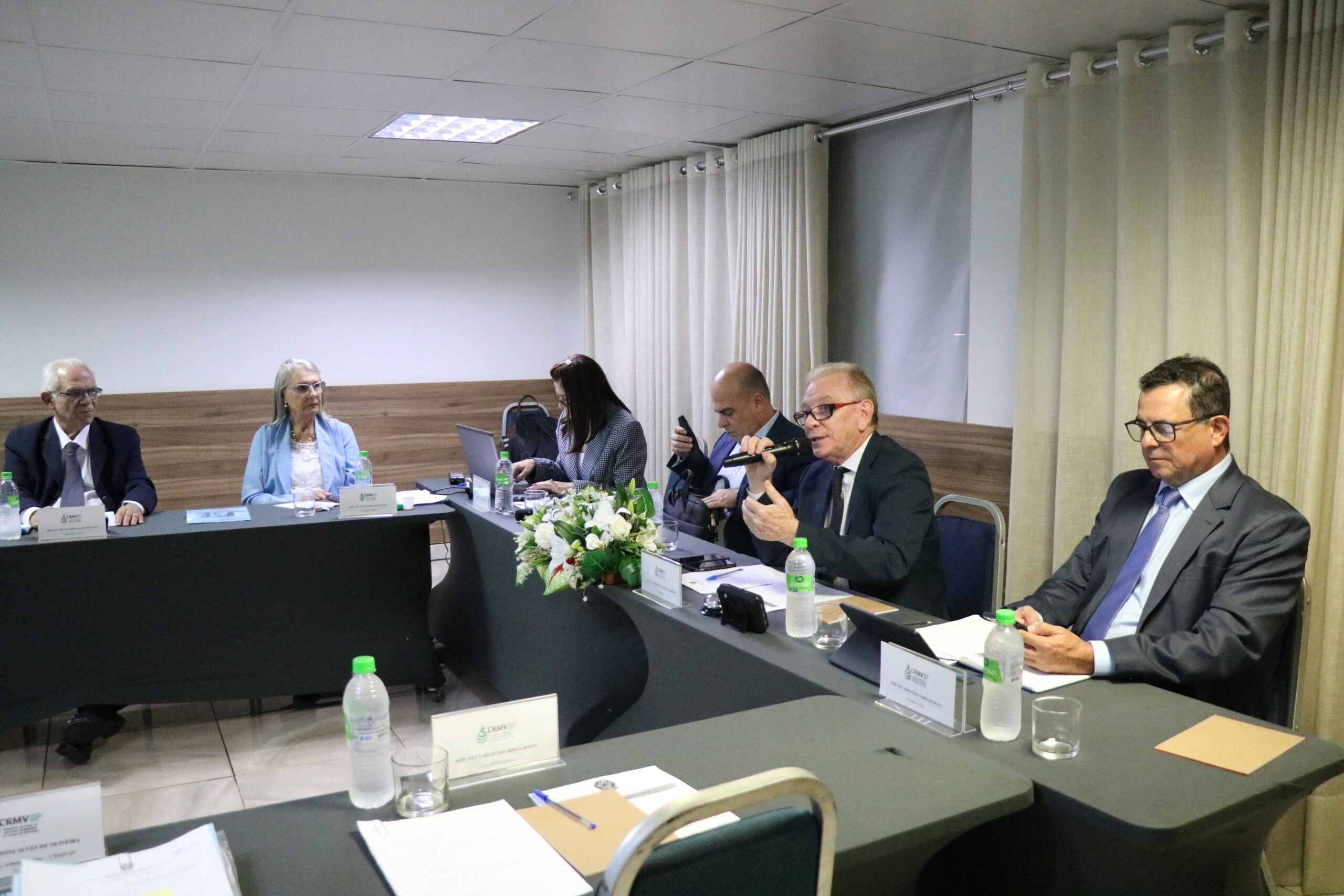 A imagem aproximada, traz seis pessoas, duas mulheres e quatro homens, sendo três deles de óculos. Eles estão sentados à mesa, um deles está falando ao microfone.