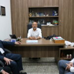 A imagem traz o vereador, de óculos e camisa branca, sentado à sua mesa, conversando com três homens de terno, que estão sentados à sua frente.