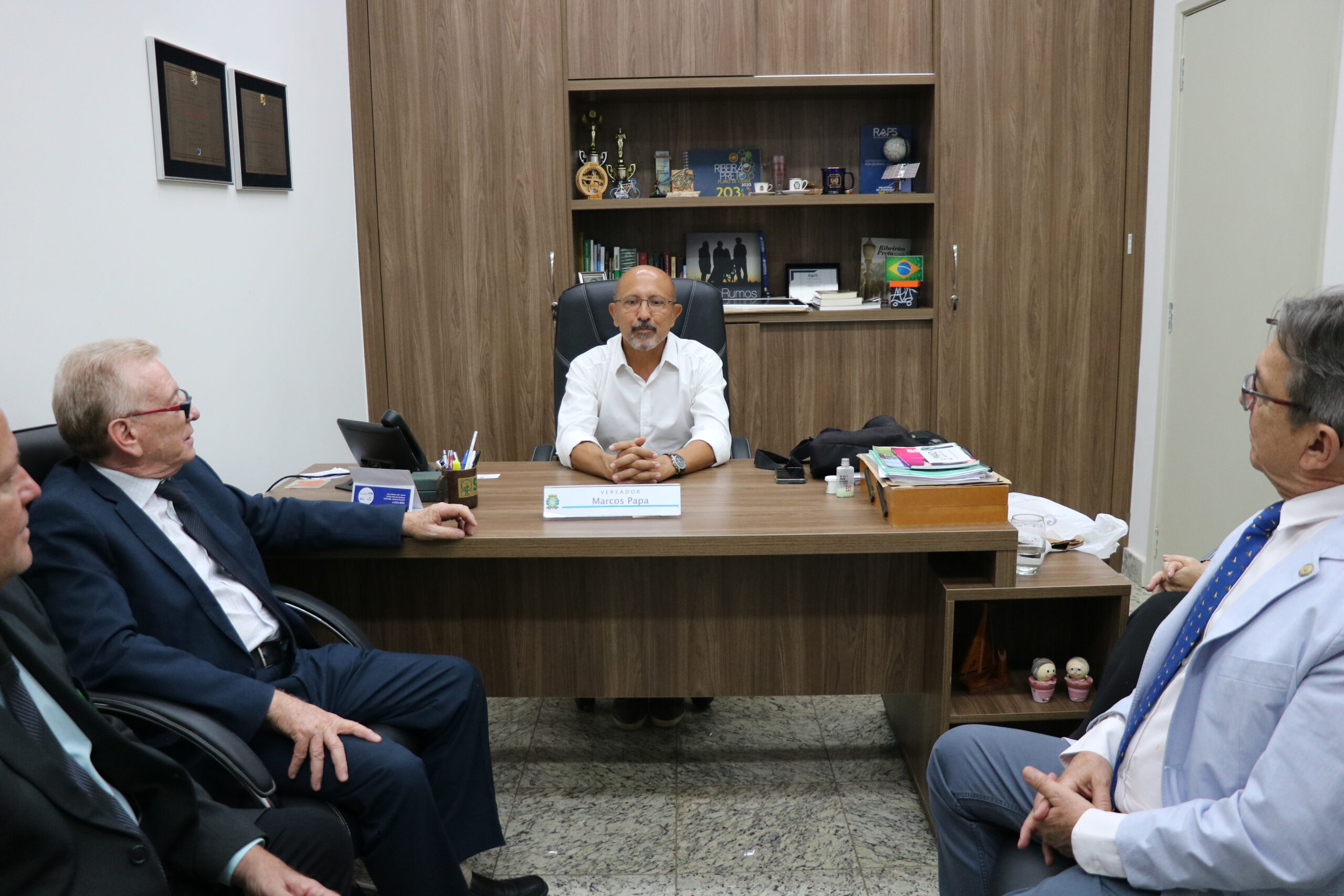 A imagem traz o vereador, de óculos e camisa branca, sentado à sua mesa, conversando com três homens de terno, que estão sentados à sua frente.