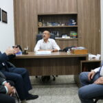 A imagem traz, no centro, um homem de óculos e camisa branca, sentado à mesa, ouvindo o que um homem à esquerda, de terno fala. Ao lado dele mais dois homens participam da conversa, ambos vestidos de terno.