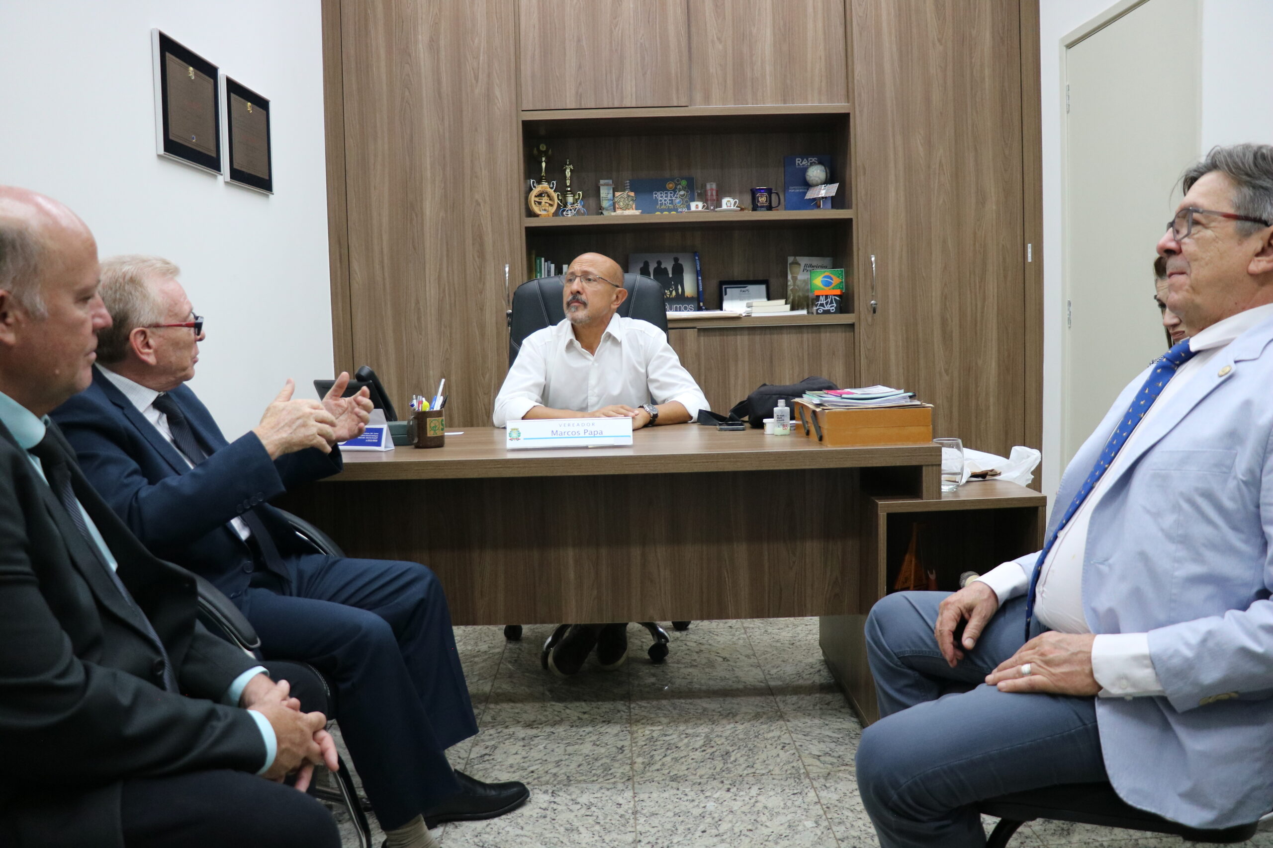 A imagem traz, no centro, um homem de óculos e camisa branca, sentado à mesa, ouvindo o que um homem à esquerda, de terno fala. Ao lado dele mais dois homens participam da conversa, ambos vestidos de terno.