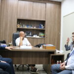 A imagem traz uma mulher de blazer xadrez, sentada em uma cadeira, em frente à mesa, onde um homem de camisa branca e óculos a ouve. Participando da reunião, mais três homens, sentados, um ao lado da mulher, e outros dois à sua frente.