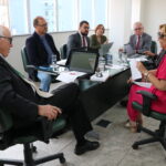 Na foto, estão sentados e conversam os participantes da reunião do PRODES: o presidente do CRMV-SP, Odemilson Donizete Mossero (primeiro ao fundo); o conselheiro do CFMV, Paulo Araújo de Guerra (à direita); a vice-presidente do CFMV, Ana Elisa de Souza Almeida; o conselheiro do CFMV, Marcílio Magalhães Vaz de Oliveira (primeiro, à frente); o presidente do CRMV-BA, Altair Santana de Oliveira (primeiro, à direita); o assessor administrativo, chefe do Núcleo de Apoio aos Regionais (NAR) do CFMV, Igor Pinto de Andrade; e a presidente do CRMV-PE, Maria Elisa Araújo.