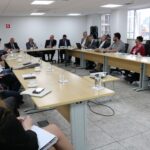 Sala ampla com mesas da cor bege dispostas em forma de "U". Ao entorno estão sentados os presidentes das comissões do CRMV-SP, entre os quais mulheres e homens. Ao fundo, a Diretoria Executiva do CRMV-SP e o presidente do CFMV. O presidente do CFMV está com a palavra e segura um microfone.