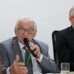 Presidente do CFMV está com a palavra. Ele é um homem branco, está sentado, de terno e gravata cinzas, usa óculos, tem cabelos brancos e segura um microfone. Ao seu lado, o presidente do CRMV-SP, homem branco, de cabelos grisalhos, com terno preto, gravata estampada e óculos de aro preto.