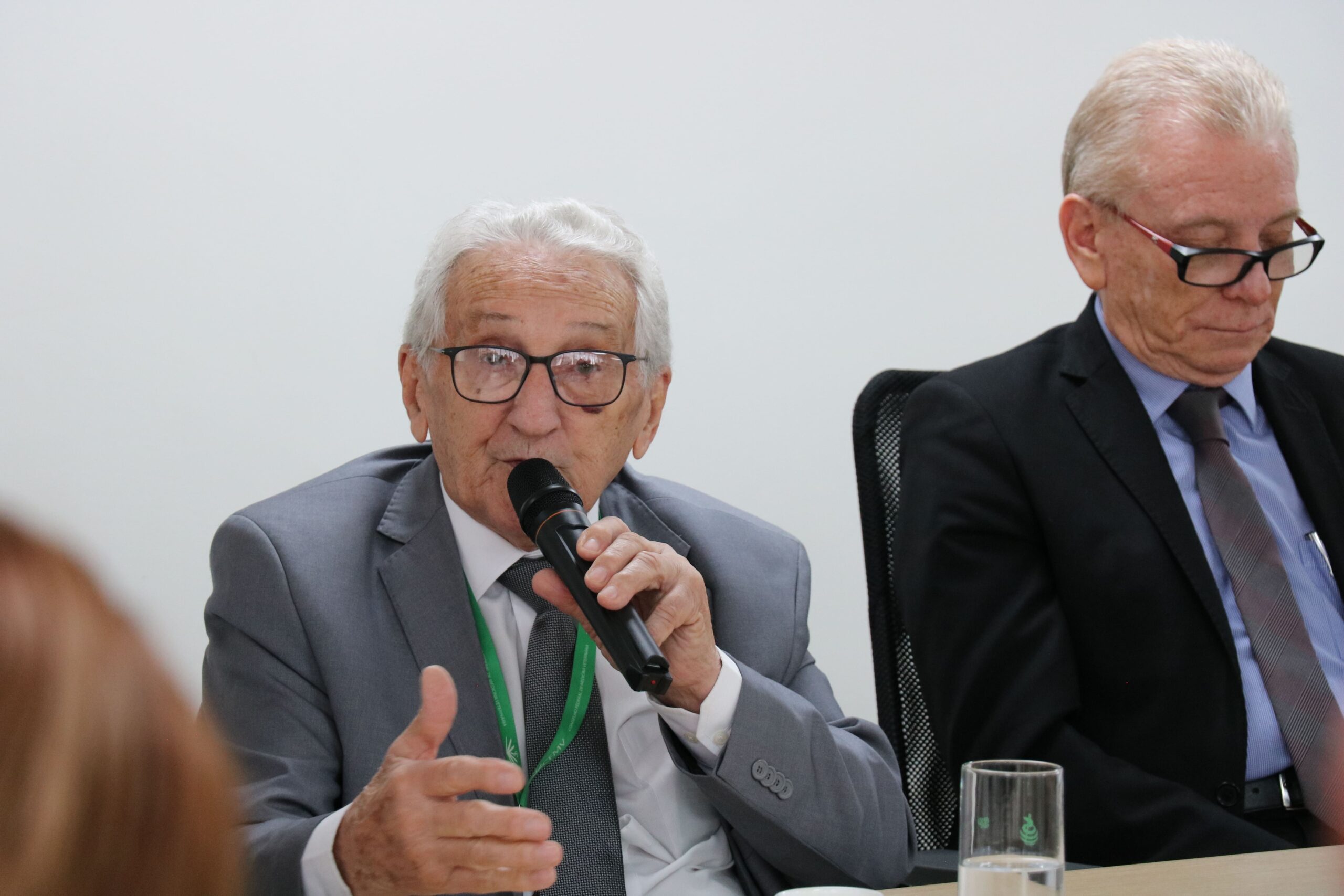 Presidente do CFMV está com a palavra. Ele é um homem branco, está sentado, de terno e gravata cinzas, usa óculos, tem cabelos brancos e segura um microfone. Ao seu lado, o presidente do CRMV-SP, homem branco, de cabelos grisalhos, com terno preto, gravata estampada e óculos de aro preto.