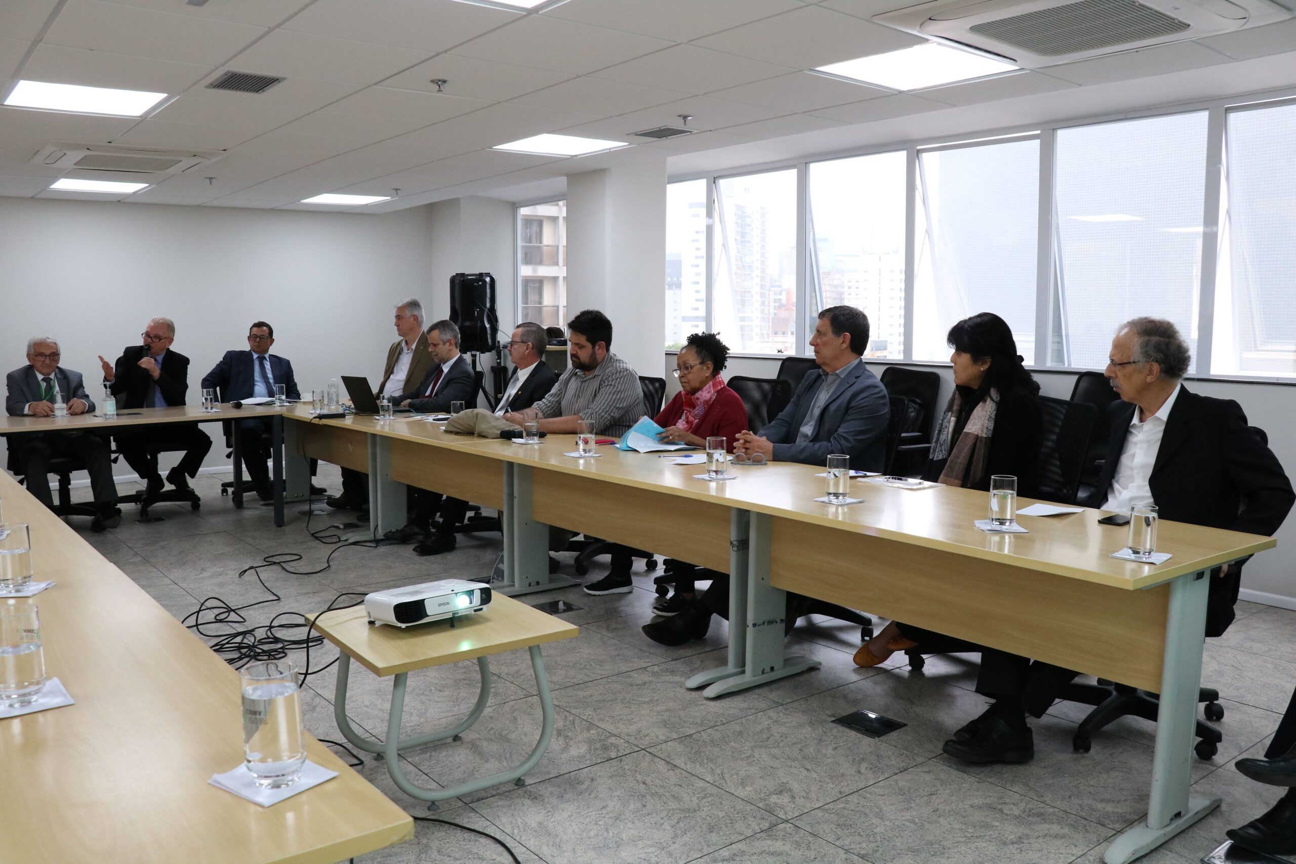 Visão lateral da sala. Em que é possível ver os presentes em dois dos lados do "U" formado pela disposição das mesas. No canto esquerdo da imagem, o presidente do CRMV-SP fala do microfone.