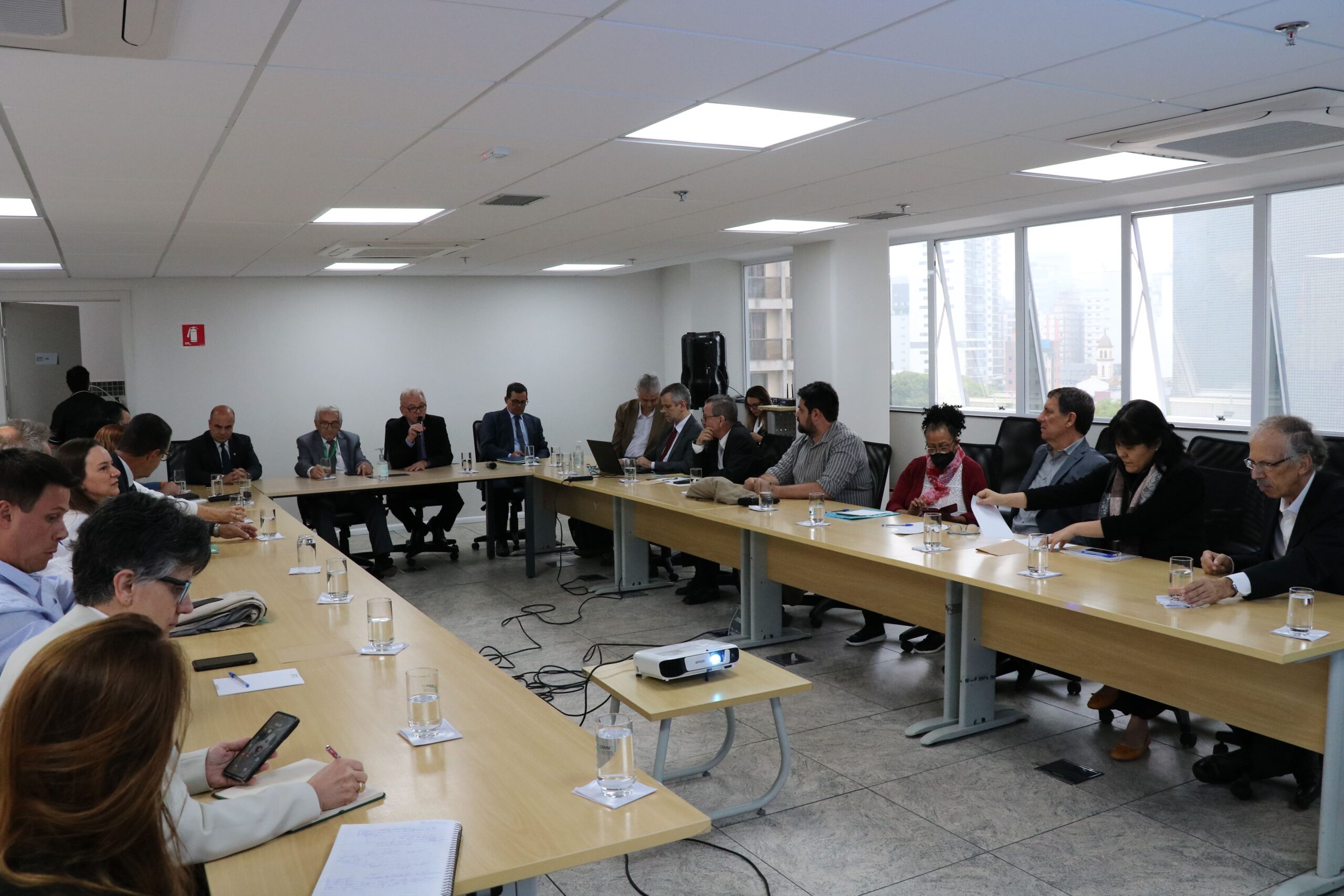 Sala ampla com mesas na cor bege dispostas em formato de "U". Ao entorno estão sentados presidentes de comissões, coordenadores e membros da diretoria do CRMV-SP, bem como o presidente do CFMV. Todos estão atentos a fala do presidente do Regional, o qual seria o microfone.