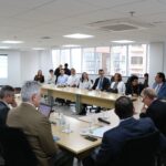 Sala ampla com mesas dispostas em formato de "U", com os presidentes das comissões sentados ao entorno. No fundo da sala um telão com projeção e uma janela ampla com vista para os prédios vizinhos. Todos estão atentos a fala do presidente do CFMV.