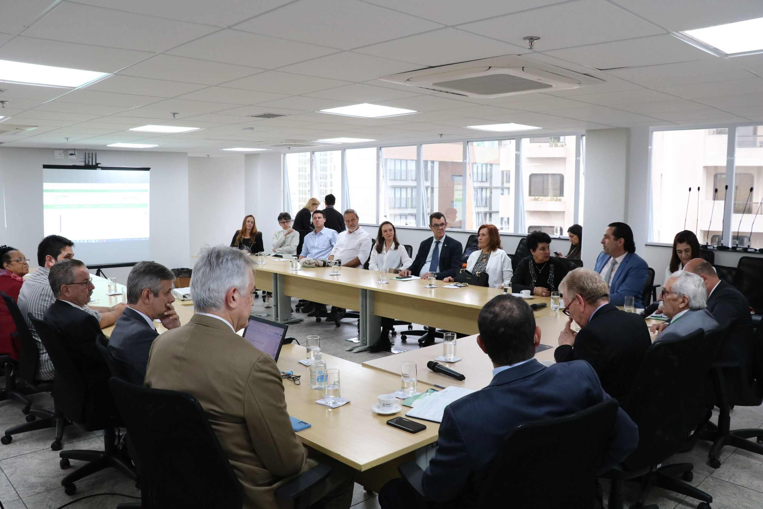 Sala ampla com mesas dispostas em formato de "U", com os presidentes das comissões sentados ao entorno. No fundo da sala um telão com projeção e uma janela ampla com vista para os prédios vizinhos. Todos estão atentos a fala do presidente do CFMV.