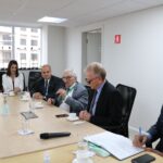 Parte da mesa em que estão os integrantes da Diretoria Executiva e o presidente do Federal, bem como o conselheiro do CRMV-SP. Todos estão sentados e acompanham atentos a fala do presidente do Federal, o qual segura um microfone. O secretário-geral do CRMV-SP está no canto direito da foto e assina alguns documentos. São cinco homens de terno e uma mulher de casaco branco.