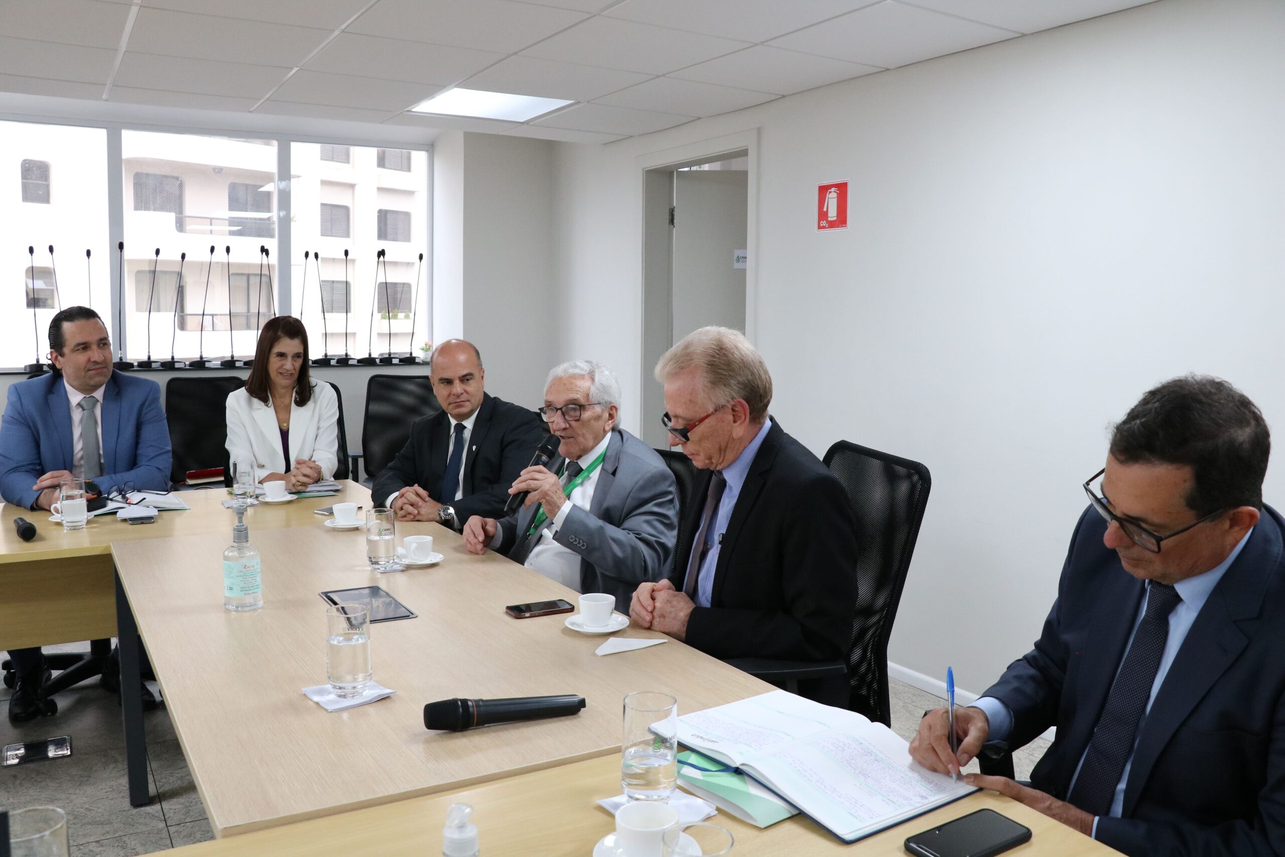 Parte da mesa em que estão os integrantes da Diretoria Executiva e o presidente do Federal, bem como o conselheiro do CRMV-SP. Todos estão sentados e acompanham atentos a fala do presidente do Federal, o qual segura um microfone. O secretário-geral do CRMV-SP está no canto direito da foto e assina alguns documentos. São cinco homens de terno e uma mulher de casaco branco.