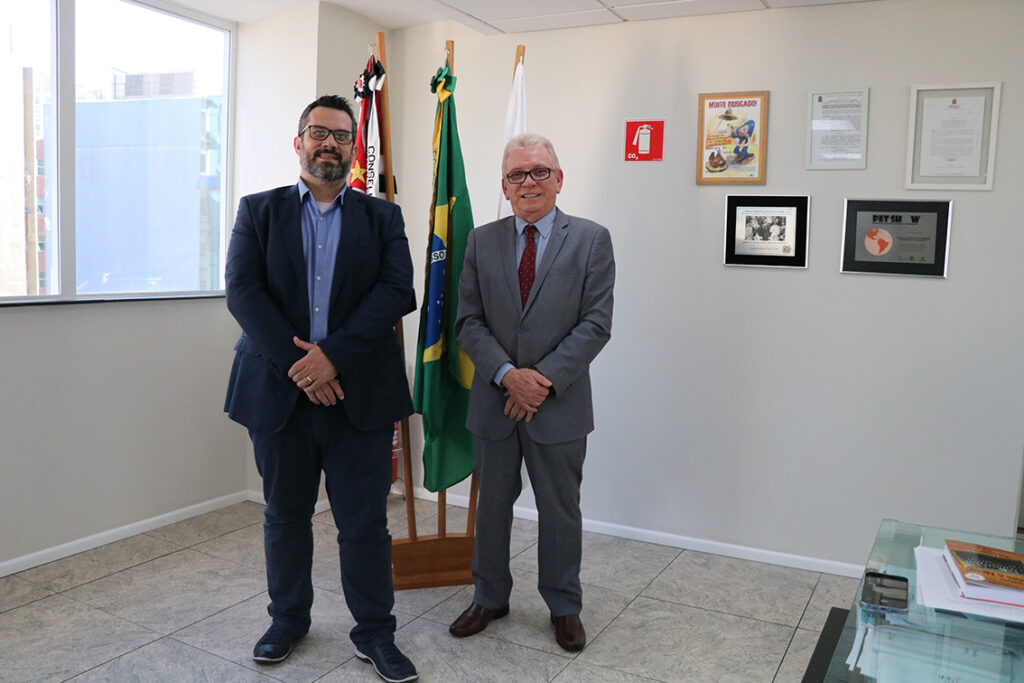 Na foto, o presidente do CRMV-SP, Odemilson Donizete Mossero, está ao lado do presidente da Comissão de Clínicos de Pequenos Animais do Regional, Márcio Thomazo Mota. Ao fundo, há bandeiras do Brasil, do estado de São Paulo e do CRMV-SP, bem como na parede há cinco quadros. Ao lado esquerdo há uma janela e ao lado direito, uma mesa.