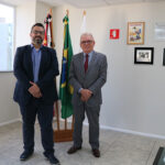 Na foto, o presidente do CRMV-SP, Odemilson Donizete Mossero, está ao lado do presidente da Comissão de Clínicos de Pequenos Animais do Regional, Márcio Thomazo Mota. Ao fundo, há bandeiras do Brasil, do estado de São Paulo e do CRMV-SP, bem como na parede há cinco quadros. Ao lado esquerdo há uma janela e ao lado direito, uma mesa.