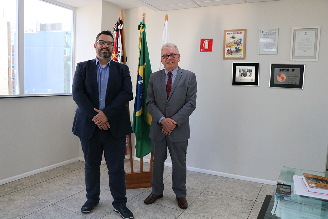 Na foto, o presidente do CRMV-SP, Odemilson Donizete Mossero, está ao lado do presidente da Comissão de Clínicos de Pequenos Animais do Regional, Márcio Thomazo Mota. Ao fundo, há bandeiras do Brasil, do estado de São Paulo e do CRMV-SP, bem como na parede há cinco quadros. Ao lado esquerdo há uma janela e ao lado direito, uma mesa.