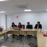 Cinco mulheres e um homem sentados à mesa, trabalhando em notebooks.