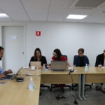 Quatro mulheres e um homem sentados à mesa de trabalho, com notebooks.