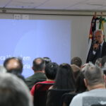 Homem de óculos, terno e gravata, com microfone na mão está no palco do auditório, falando para a plateia, formada de homens e mulheres.