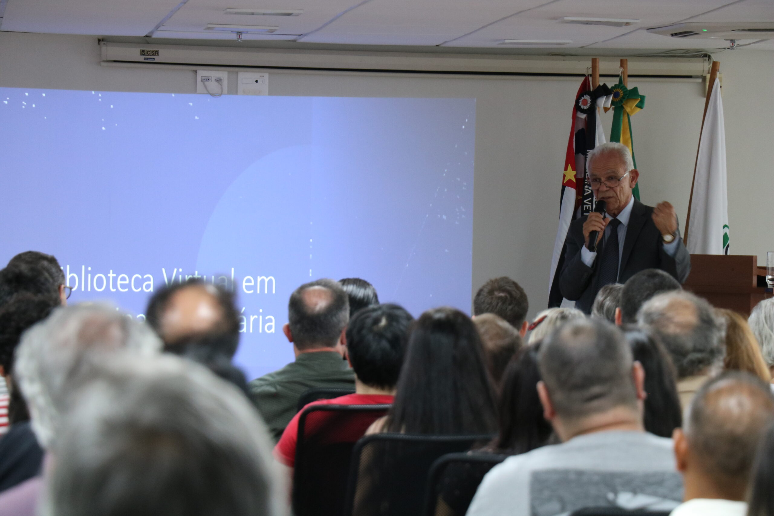 Homem de óculos, terno e gravata, com microfone na mão está no palco do auditório, falando para a plateia, formada de homens e mulheres.