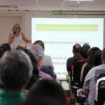 Mulher, de óculos, com vestido de alças, microfone na mão, está no palco do auditório falando para a plateia formada por homens e mulheres.