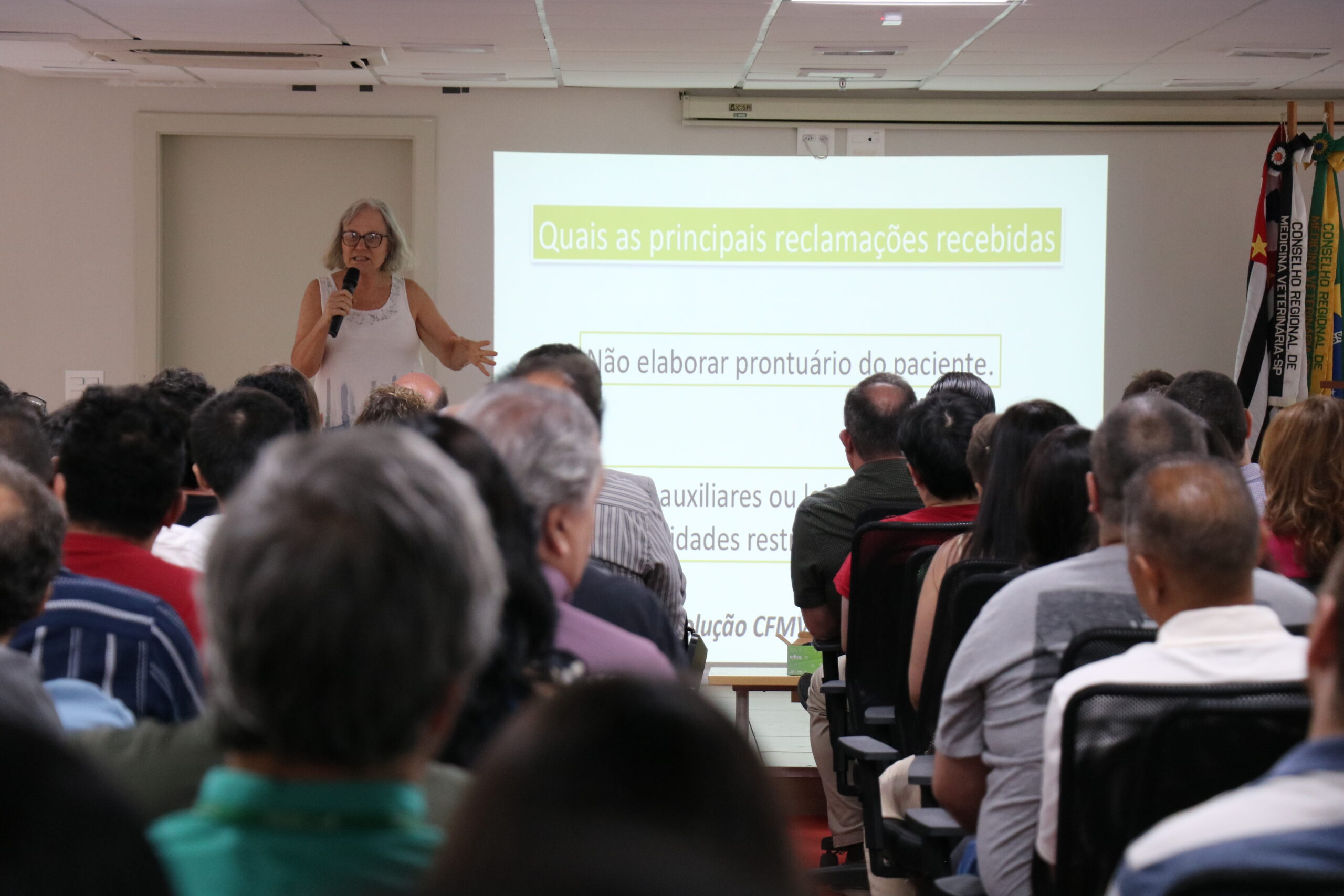 Mulher, de óculos, com vestido de alças, microfone na mão, está no palco do auditório falando para a plateia formada por homens e mulheres.