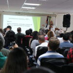 Mulher de óculos, blazer branco, microfone na mão, está no palco do auditório, com um telão ao fundo, apresentando dados para a plateia, formada por homens e mulheres.