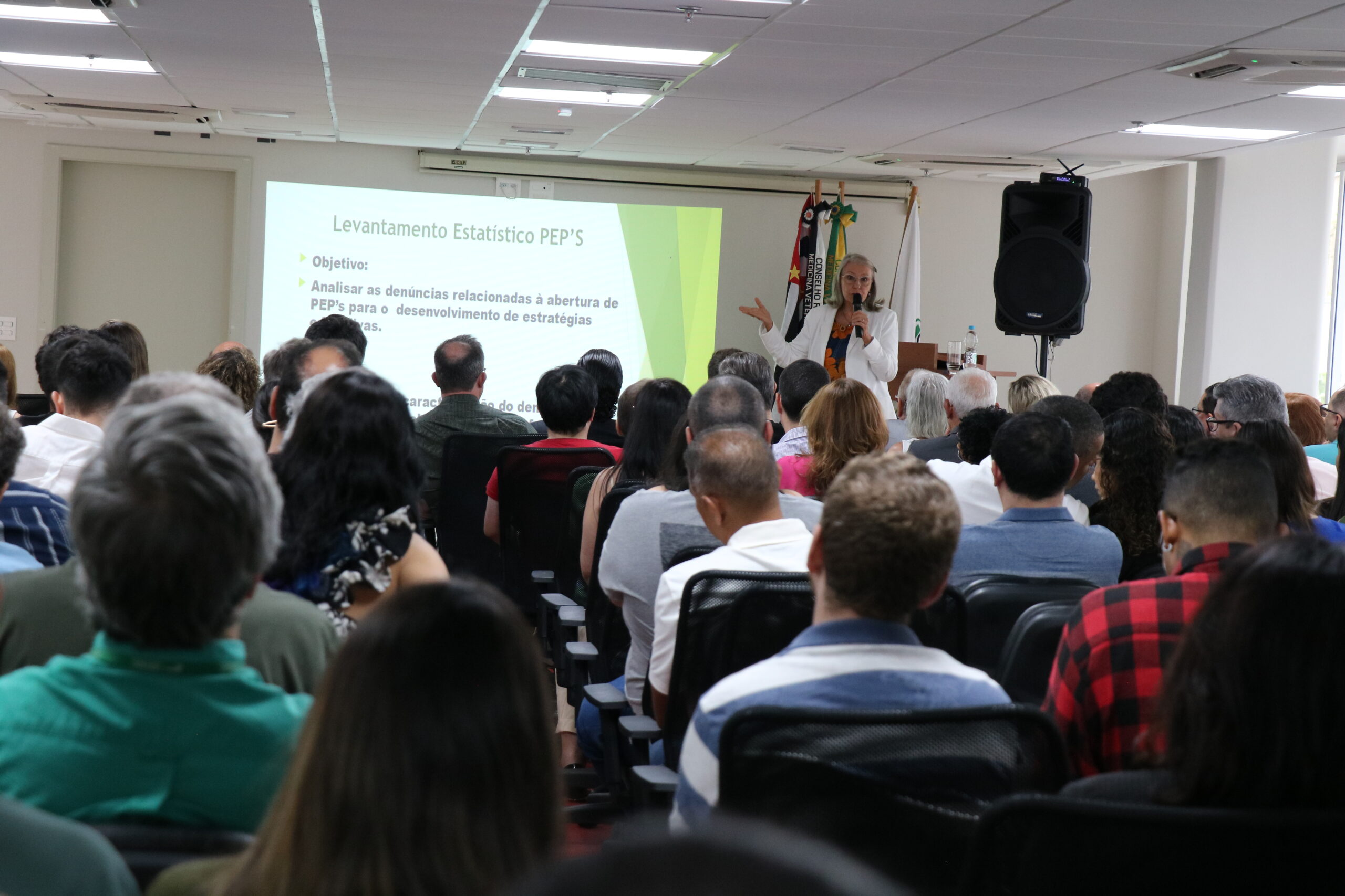 Mulher de óculos, blazer branco, microfone na mão, está no palco do auditório, com um telão ao fundo, apresentando dados para a plateia, formada por homens e mulheres.
