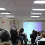 Homem, de camisa social, microfone na mão, no palco do auditório, fala para a plateia formada por homens e mulheres.