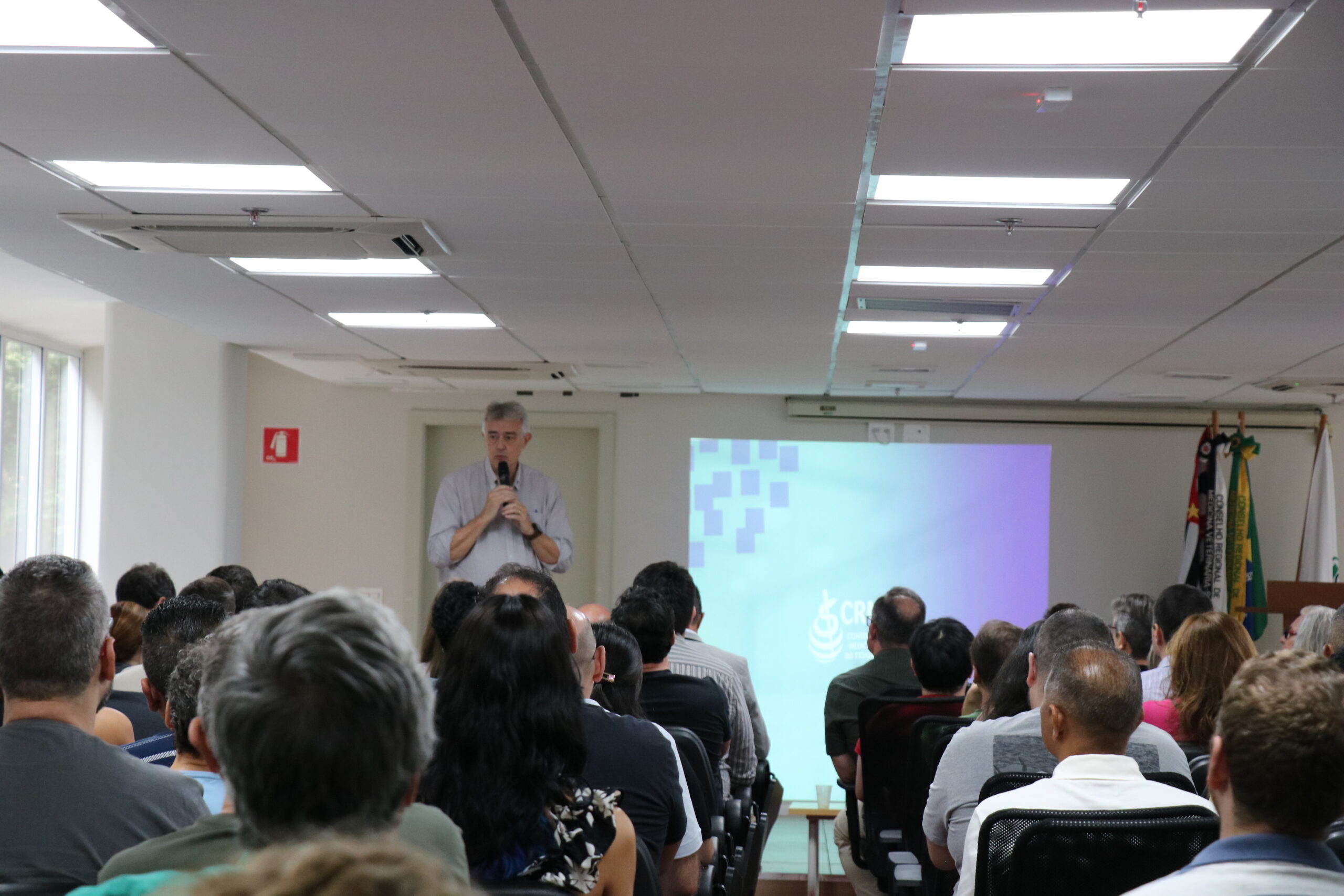Homem, de camisa social, microfone na mão, no palco do auditório, fala para a plateia formada por homens e mulheres.