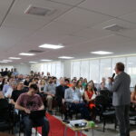 Plateia de auditório lotada, homens e mulheres sentados, algumas pessoas em pé ao fundo assistem a apresentação de um homem de terno.