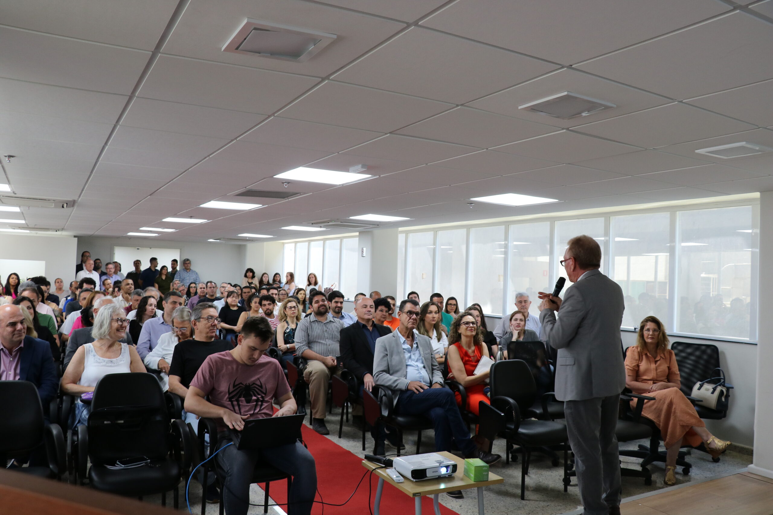 Plateia de auditório lotada, homens e mulheres sentados, algumas pessoas em pé ao fundo assistem a apresentação de um homem de terno.