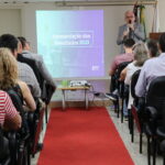 Um homem de terno e gravata, de microfone na mão, está no palco do auditório em frente a um telão fazendo a apresentação de dados para a plateia, formada de homens e mulheres, todos sentados olhando em direção ao palco.