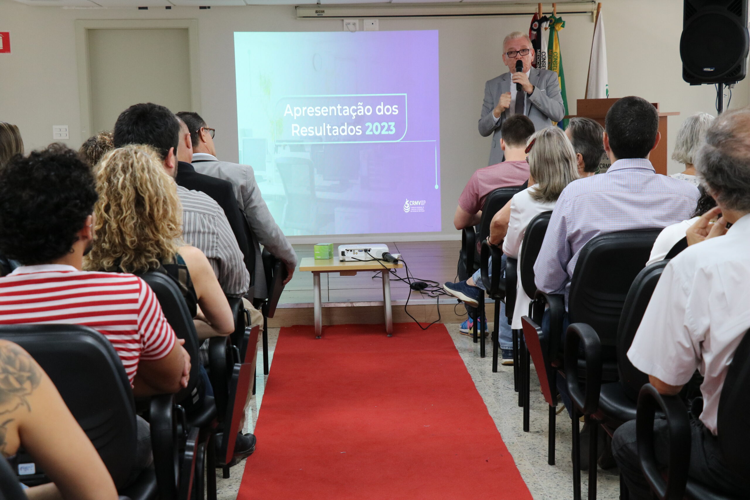 Um homem de terno e gravata, de microfone na mão, está no palco do auditório em frente a um telão fazendo a apresentação de dados para a plateia, formada de homens e mulheres, todos sentados olhando em direção ao palco.