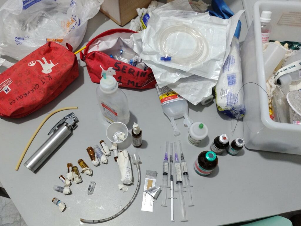 Mesa com ampolas abertas, seringas agulhadas com conteúdo não identificado, medicações de uso controlado, e equipamentos de uso cirúrgico.
