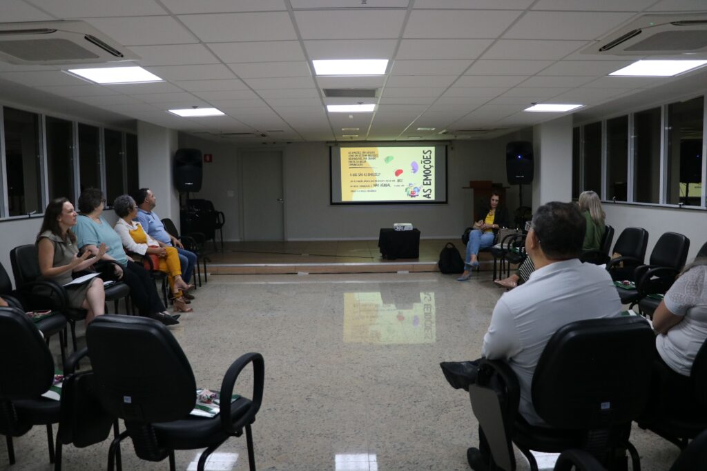 Na foto, sessão do Grupo de Apoio Mútuo. Os participantes estão sentados em cadeiras em círculos. Ao fundo, telão com explicação sobre as emoções.
