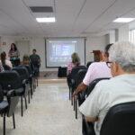 Na foto, aparecem colaboradores do CRMV-SP durante treinamento para as eleições do órgão. Eles estão sentados e aparecem de costas, viradas para a apresentadora, que está ao lado de um telão com a apresentação.