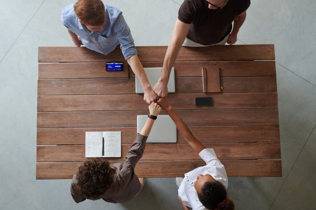 A imagem mostra quatro pessoas juntando as mãos, representando a união, em torno de uma mesa retangular de madeira.