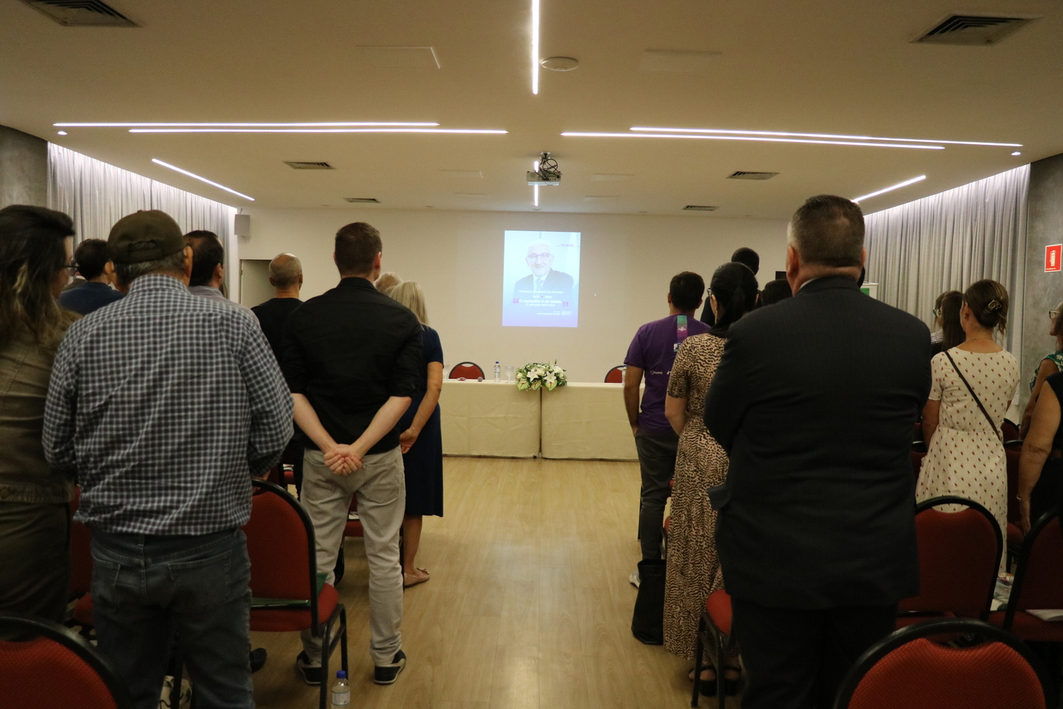 No auditório, os participantes do evento se colocam de pé, respeitam um minuto de silêncio em memória do ex-presidente do CFMV, cuja imagem aparece no telão ao fundo.