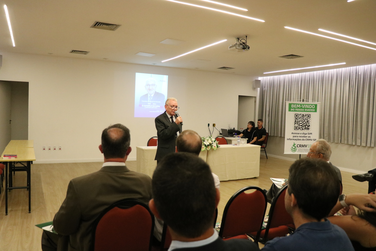 O presidente do CRMV-SP, durante homenagem ao ex-presidente do CFMV, falecido recentemente, fala ao microfone para a plateia, com a imagem do homenageado no telão ao fundo.