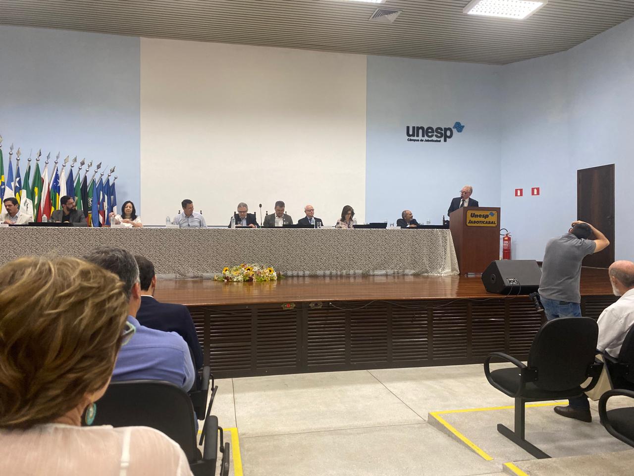 Presidente do CRMV-SP, Odemilson Donizete Mossero, está discursando aos presentes no quinquagésimo aniversário do hospital veterinário da Unesp. Ele está em um púlpito e ao lado há uma mesa em que sentam as outras autoridades presentes na solenidade.