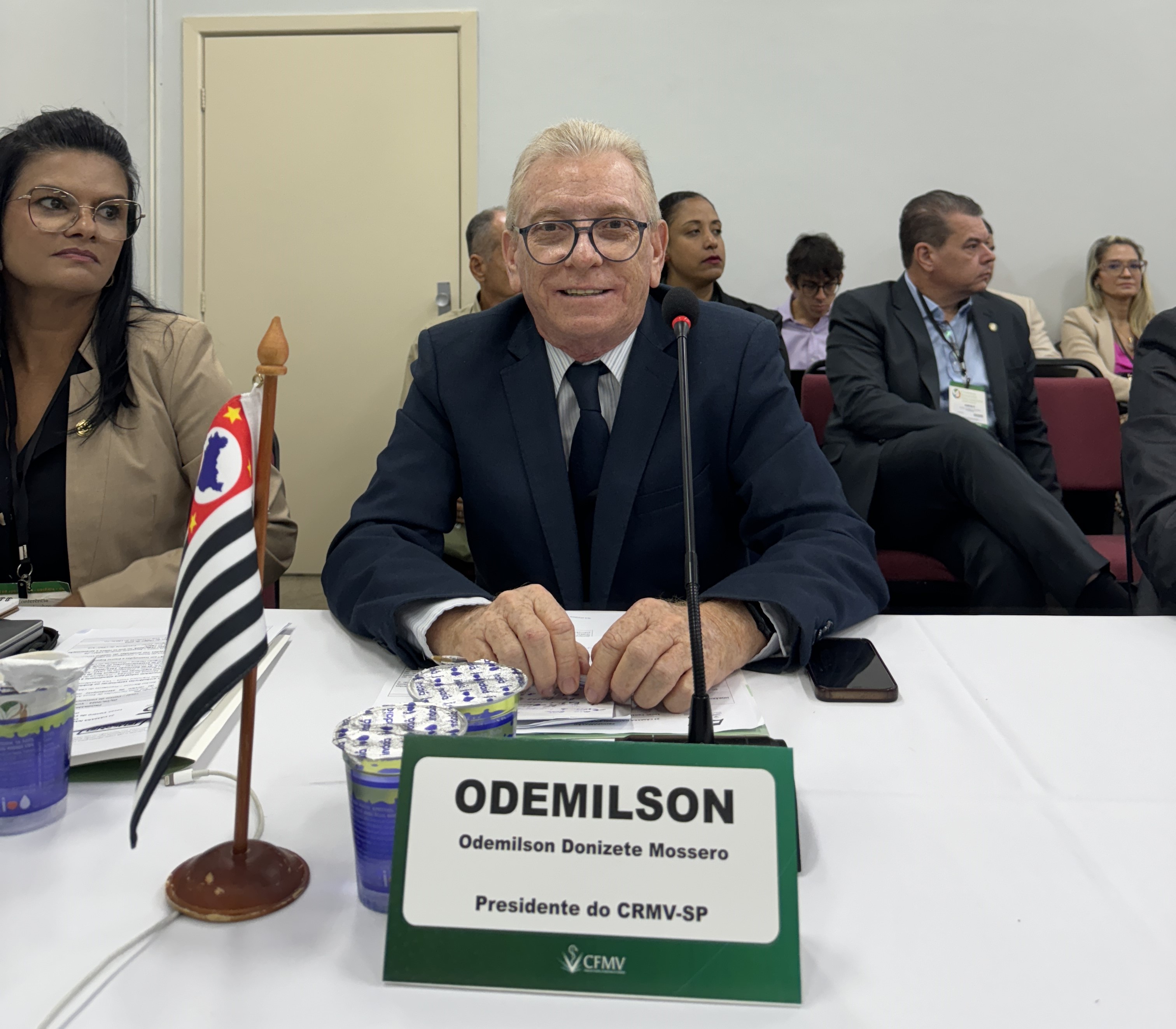 O presidente do Conselho, Odemilson Donizete Mossero, sentado à mesa, que tem uma placa de identificação com seu nome e uma bandeira do estado de São Paulo.