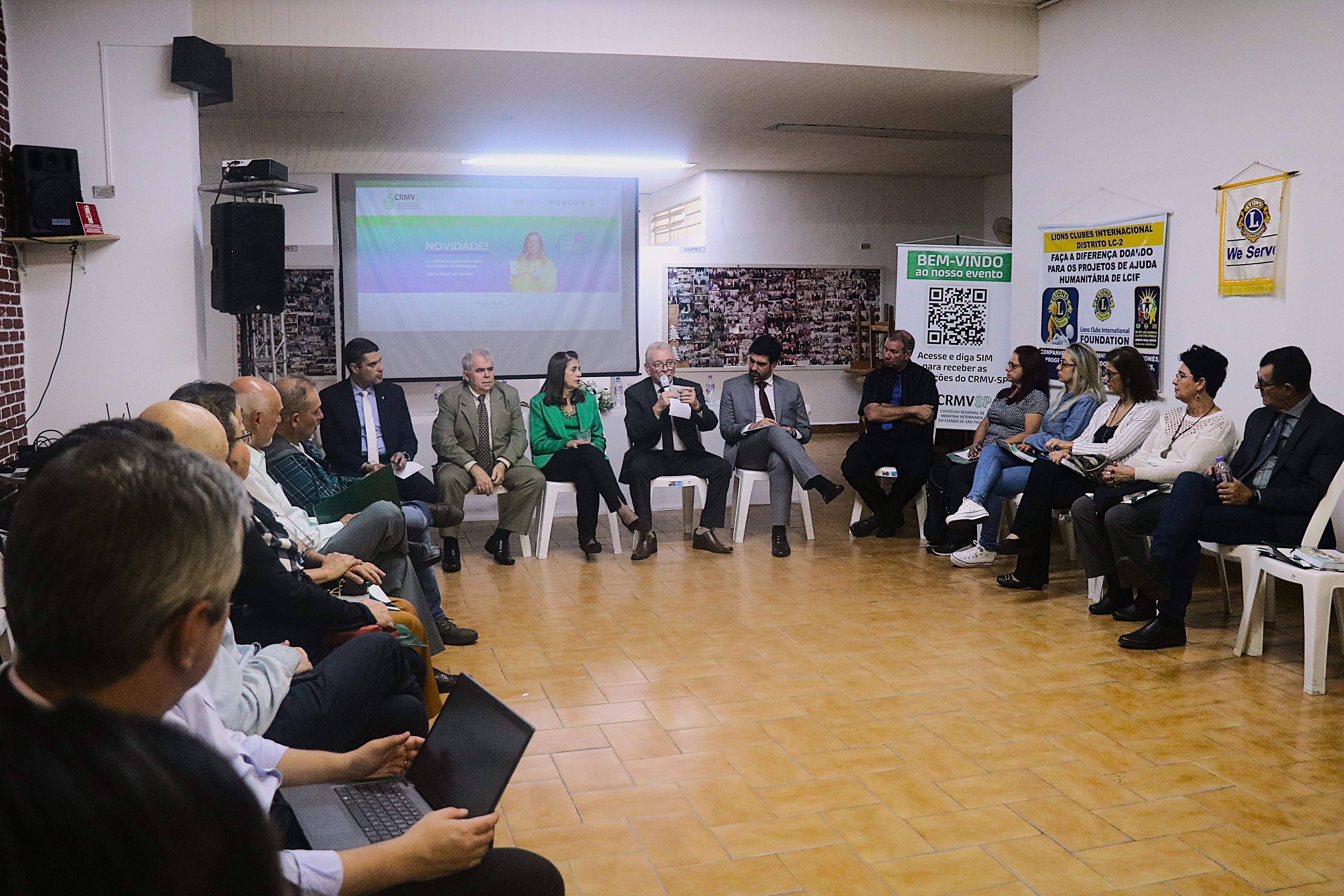 Homens e mulheres, sentados em cadeiras, organizadas em formato de U, ouvem o presidente do CRMV-SP falar ao microfone no auditório do evento.